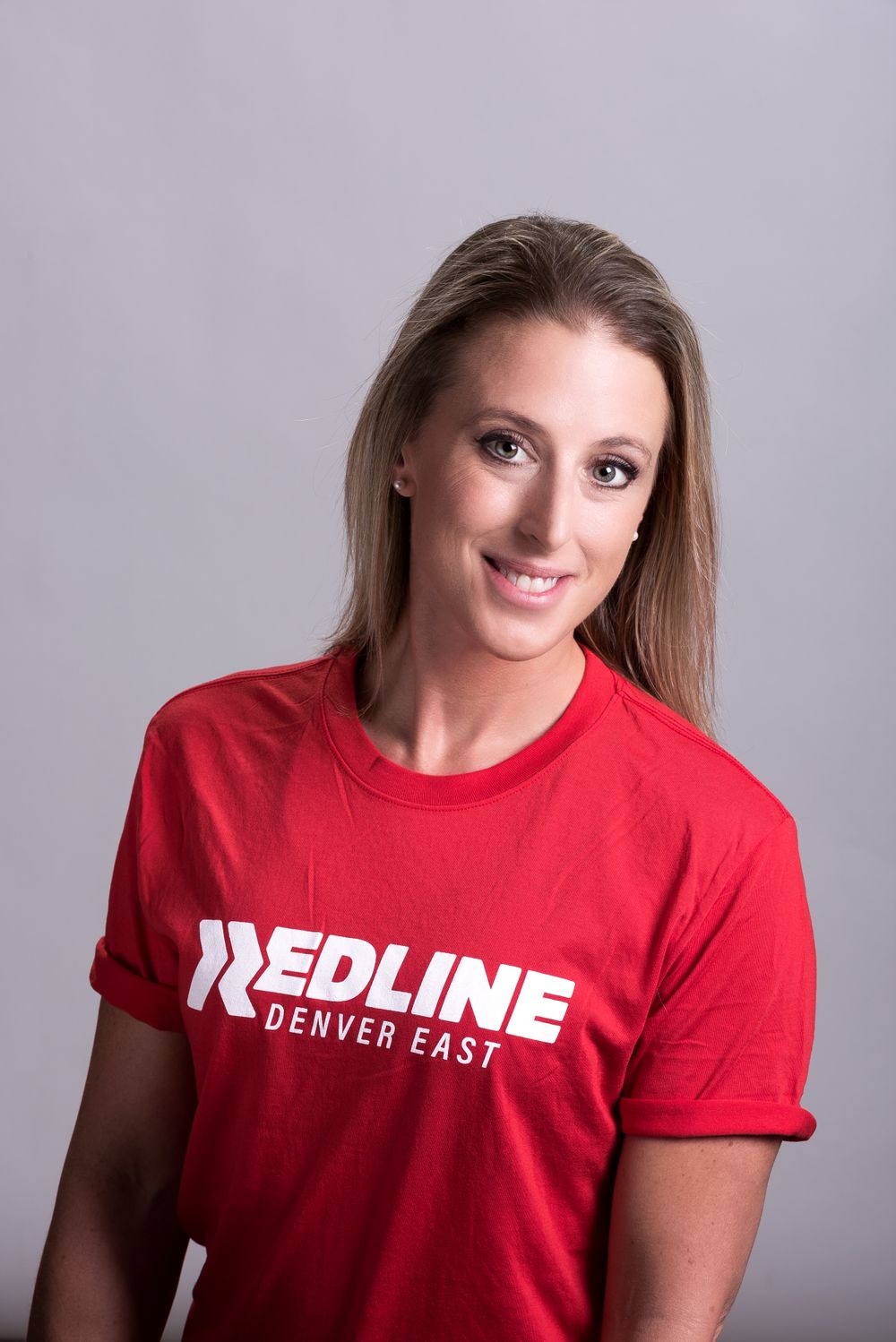 Woman in red Redline Denver East t-shirt poses for headshot against gray background.