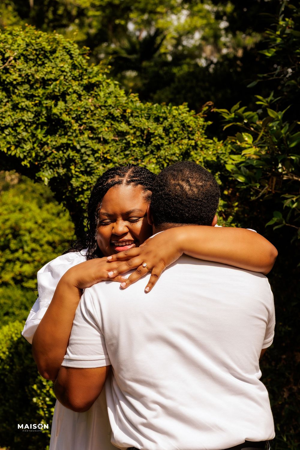 Two people sharing an intimate emotional embrace outdoors with lush green foliage in the background.