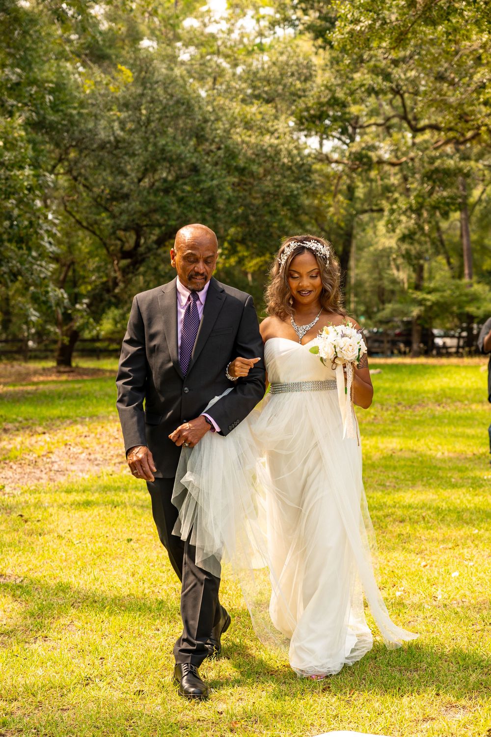 A bride in a flowing white dress walks arm-in-arm with someone in a dark suit through a sunlit park on a wedding day.