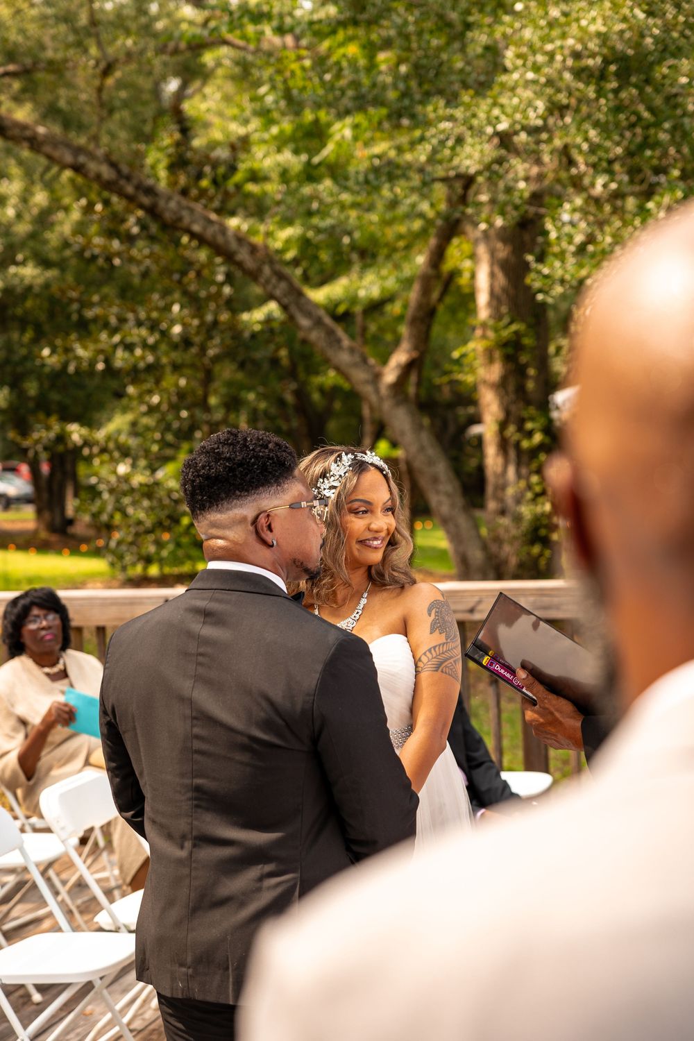 Newlyweds share a joyful moment during their outdoor wedding ceremony with guests and trees in the background.