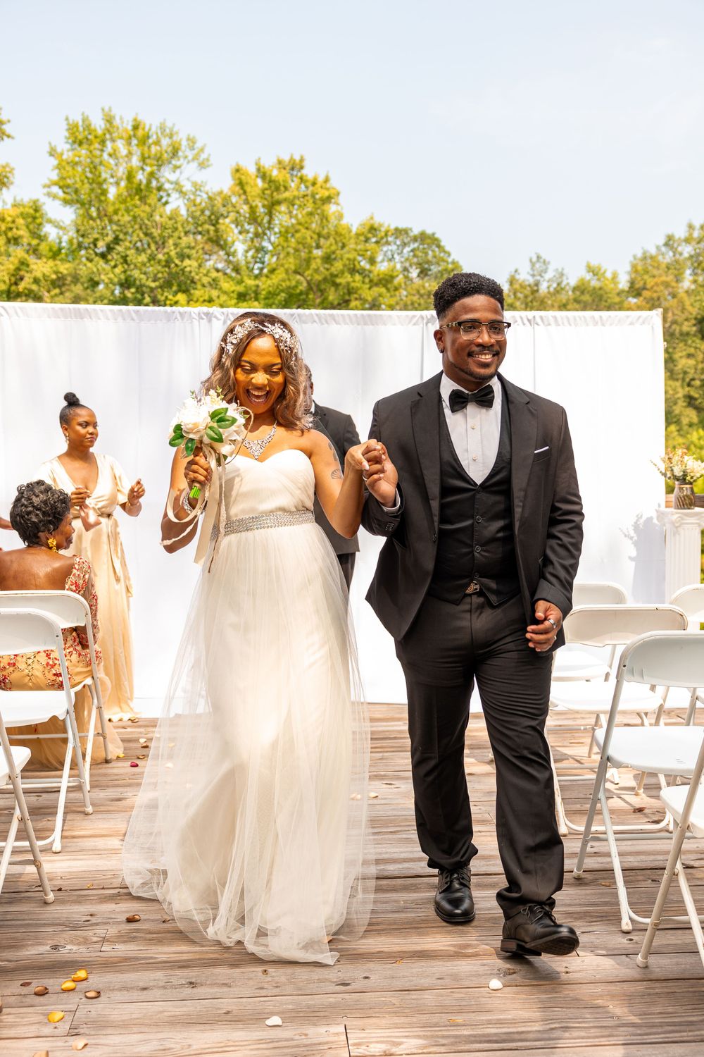 Newlyweds walking down the aisle after ceremony with white chairs and green trees in background. Engagement photographer. Wedding photograph