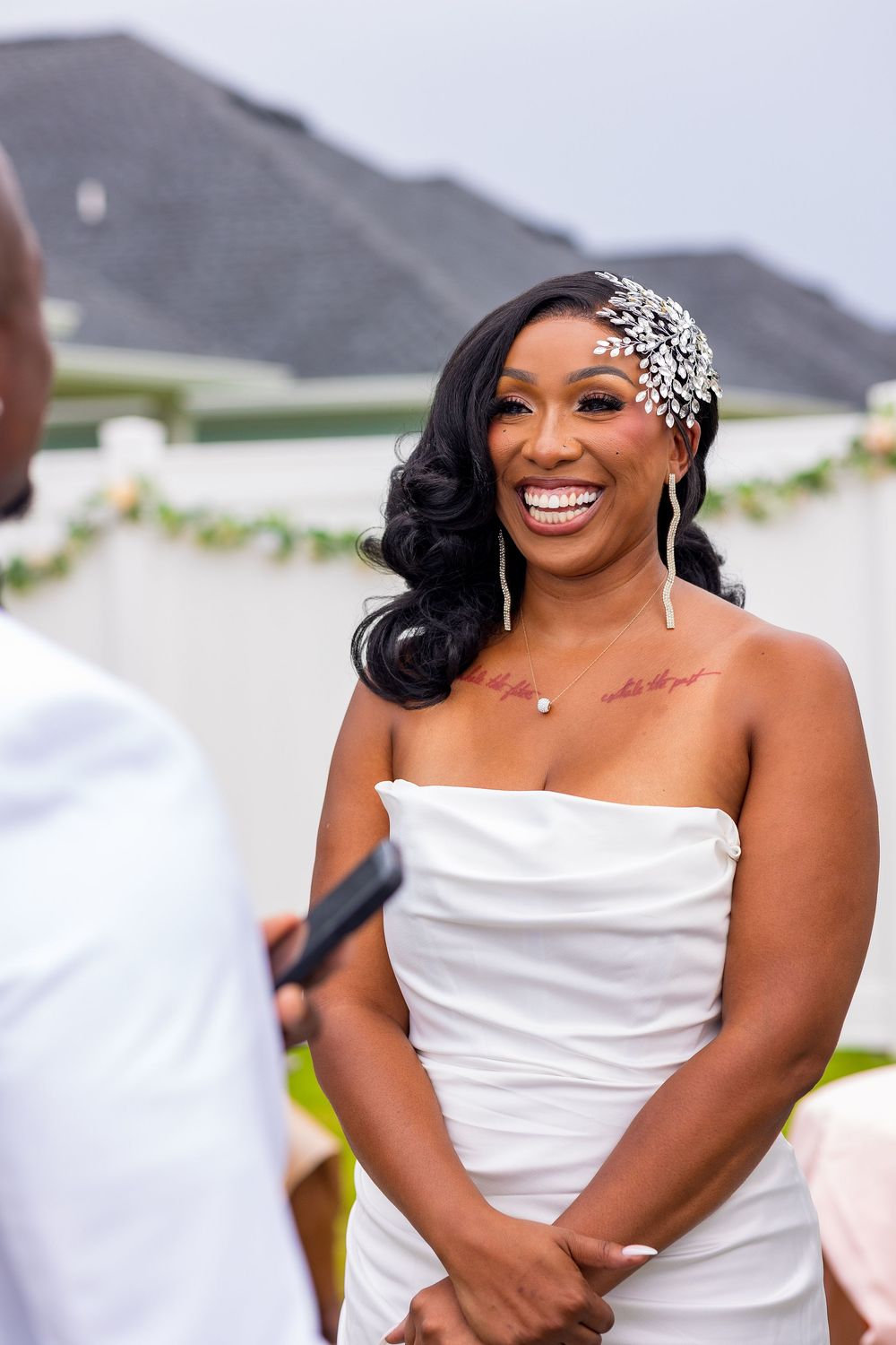 Radiant bride in white strapless gown and jeweled headpiece smiling joyfully during outdoor wedding ceremony.