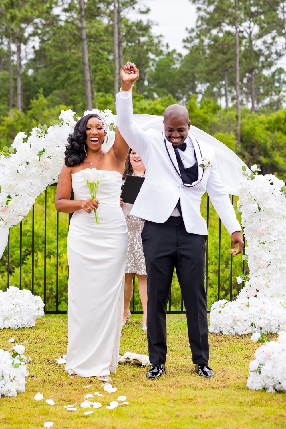 Joyful newlywed couple celebrating after outdoor wedding ceremony with white floral arch and rose petals.