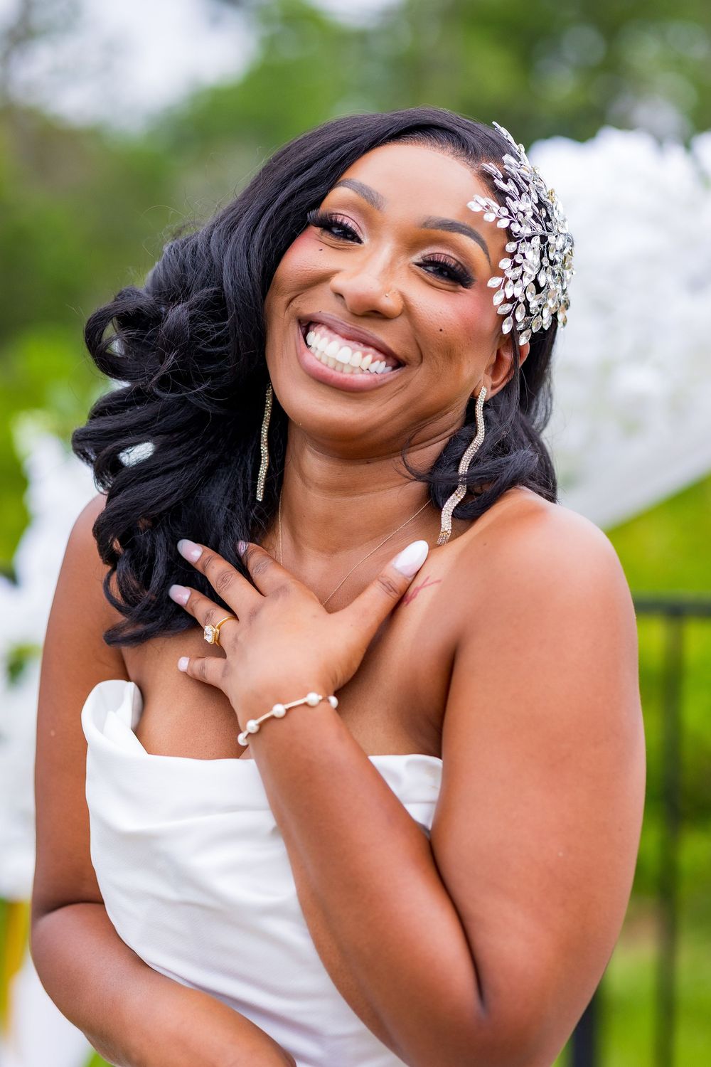 Smiling Black bride in white strapless gown with crystal hair accessory and pearl bracelet posing outdoors.
