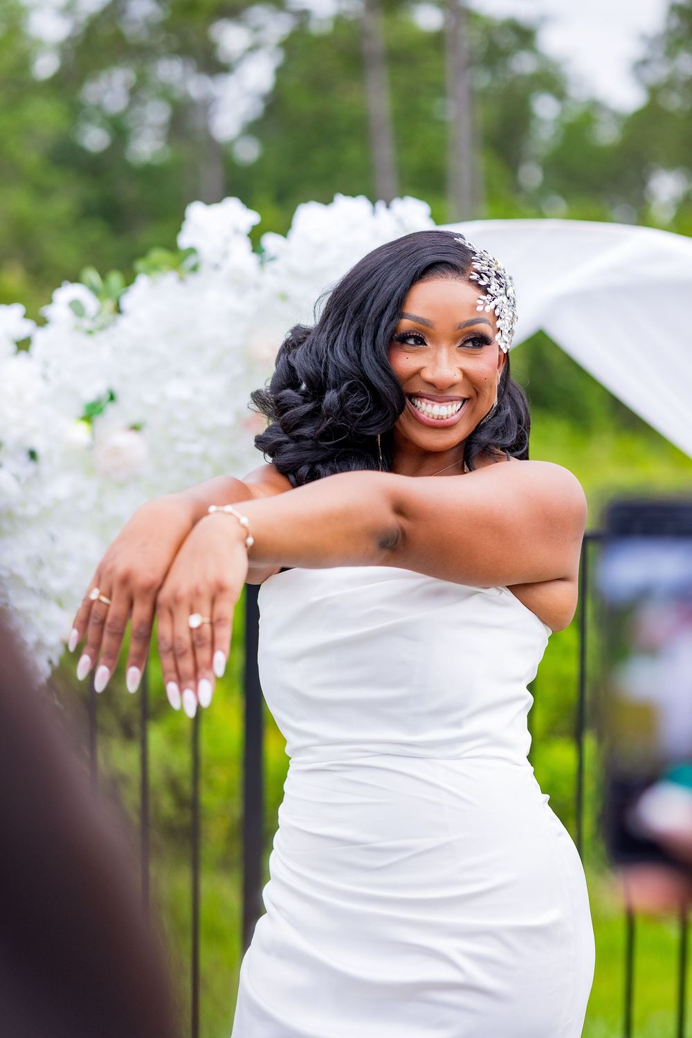 Smiling Black bride in white strapless gown with jeweled hair accessory poses outdoors at elegant garden wedding ceremony.