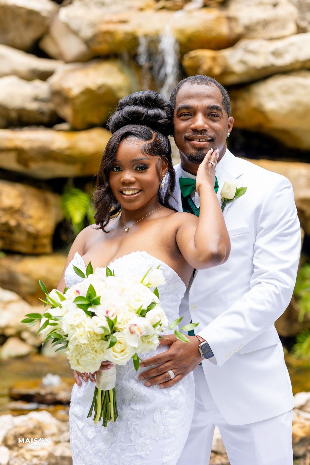 A couple poses for their wedding portrait in front of a natural stone waterfall backdrop with a white floral bouquet.