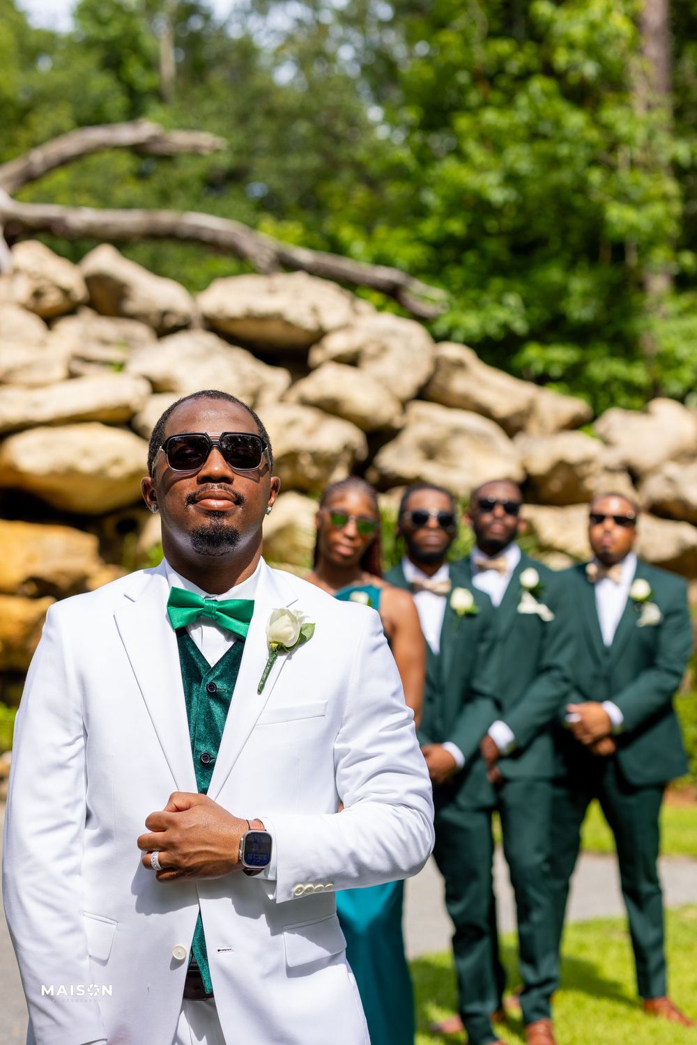 Wedding party members pose outdoors with the groom in white tuxedo and emerald green bow tie in the foreground.
