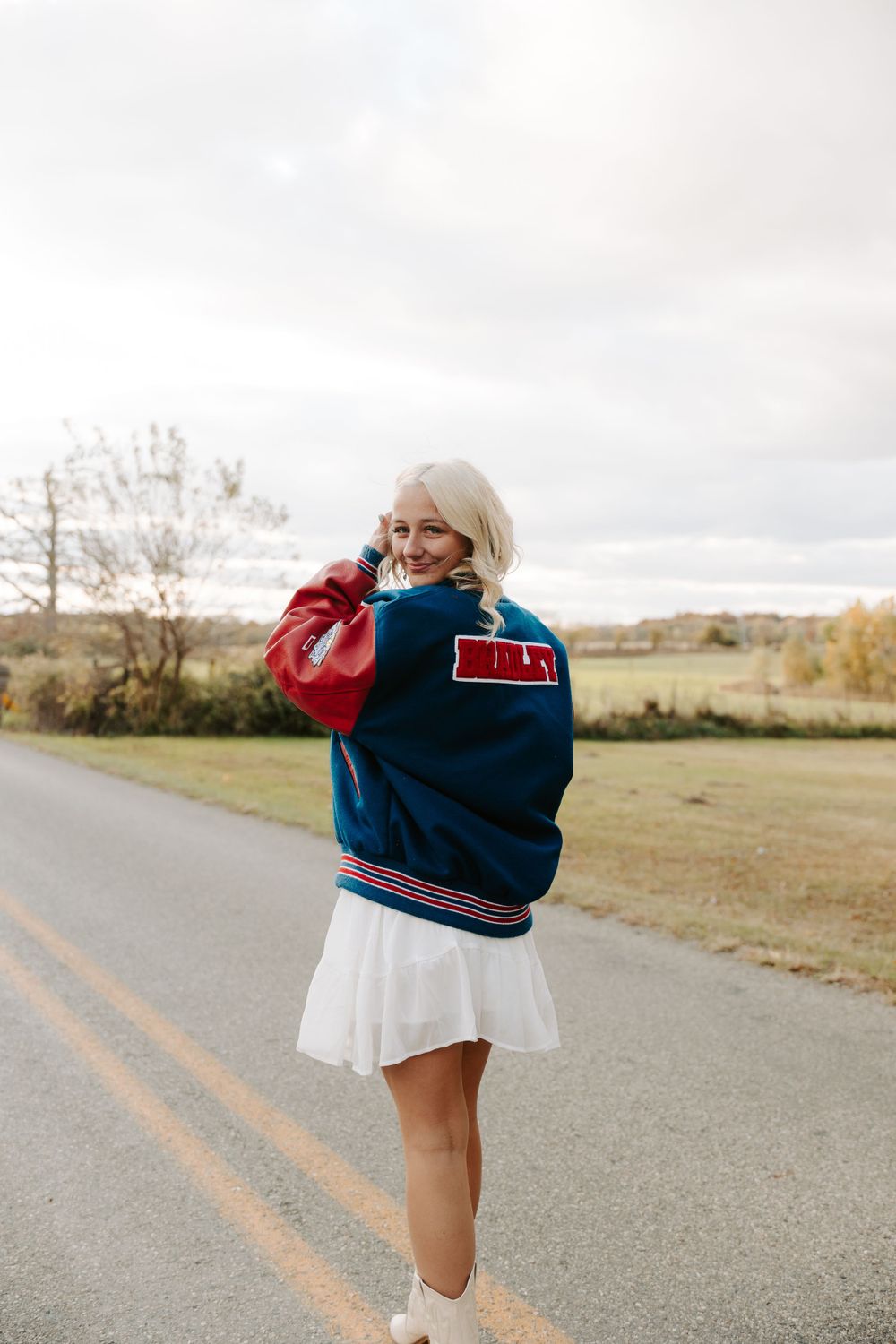 Person in vintage varsity jacket and white dress poses on country road during fall season.
