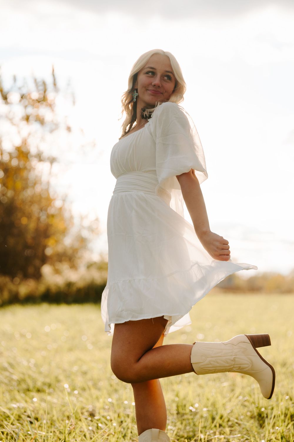 Person in white dress and boots poses playfully in sunny field during golden hour.