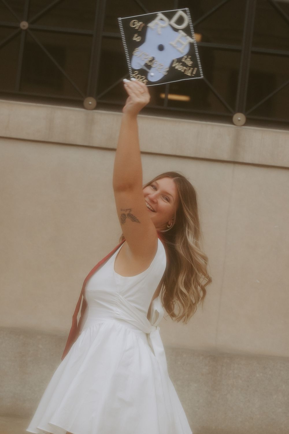 A graduate in a white dress holds up their diploma while celebrating outdoors on graduation day.