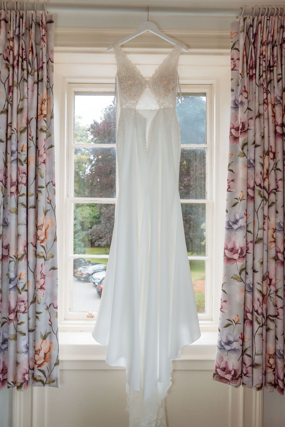 Three white wedding dresses hanging from windows in a room with floral curtains and natural lighting.