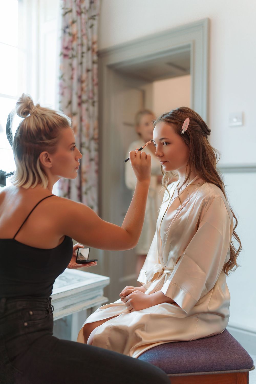 Makeup artist applying cosmetics to a client sitting on a stool in a bright room with floral curtains.