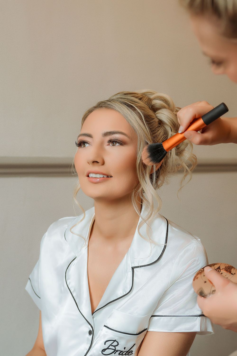 Makeup artist applies cosmetics to a person wearing a white satin robe with black piping during bridal preparation.