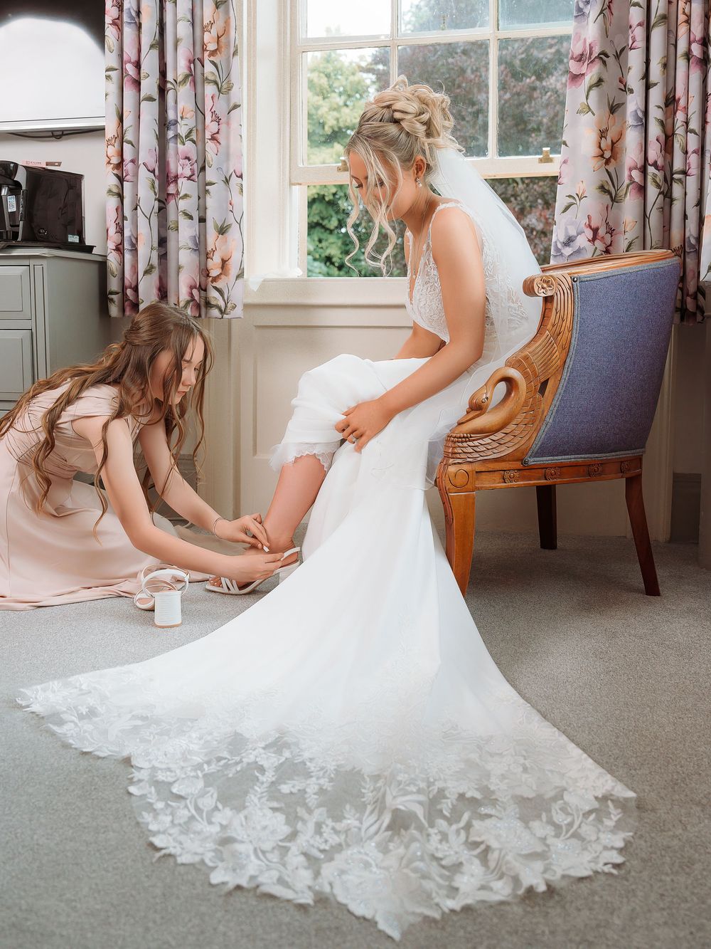 A person in a white wedding gown adjusts their shoes while sitting in an elegant room with floral curtains.