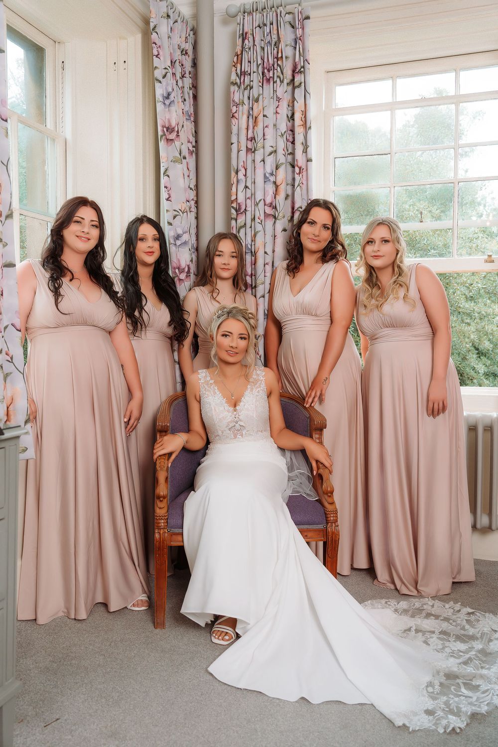 Bridal party poses in matching blush pink dresses alongside bride in white gown in bright sunlit room with floral curtains.