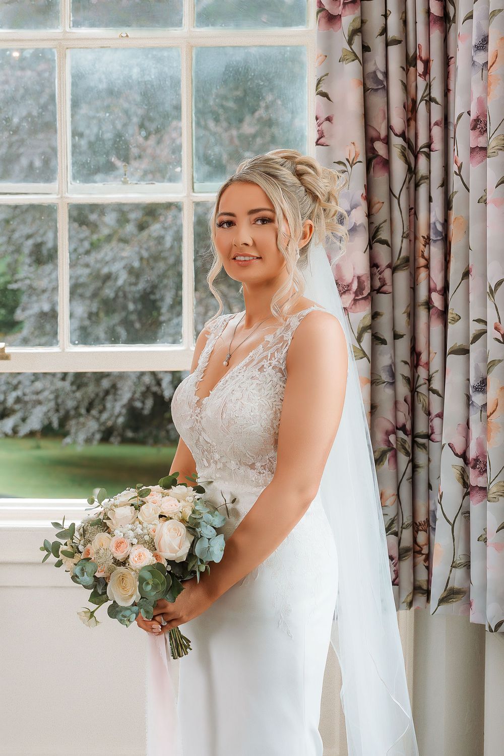 A bride in an elegant lace wedding dress poses by a window with floral curtains while holding a pastel bouquet.