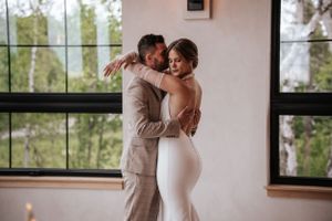 Couple embracing by large windows in elegant wedding attire during intimate moment.