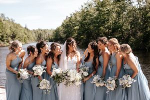 Wedding party in light blue bridesmaid dresses pose with bouquets by a lake.