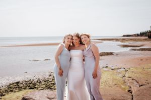 Three people in white dresses pose together on a scenic beach at sunset.
