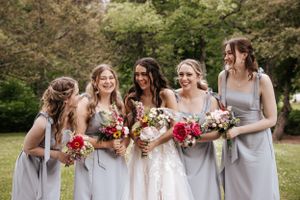 A bridal party in light gray dresses holding colorful bouquets poses outdoors.