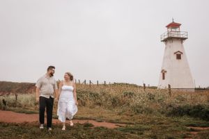 A couple walks along a red dirt path towards a white lighthouse on a cloudy day.