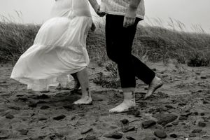 Black and white photo of barefoot couple dancing together on a beach with flowing dress and rolled up pants.