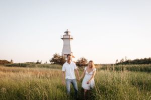 Couple poses in front of a white lighthouse surrounded by tall grass at sunset.
