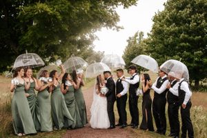 Wedding party holding clear umbrellas poses together on path in lush green setting.