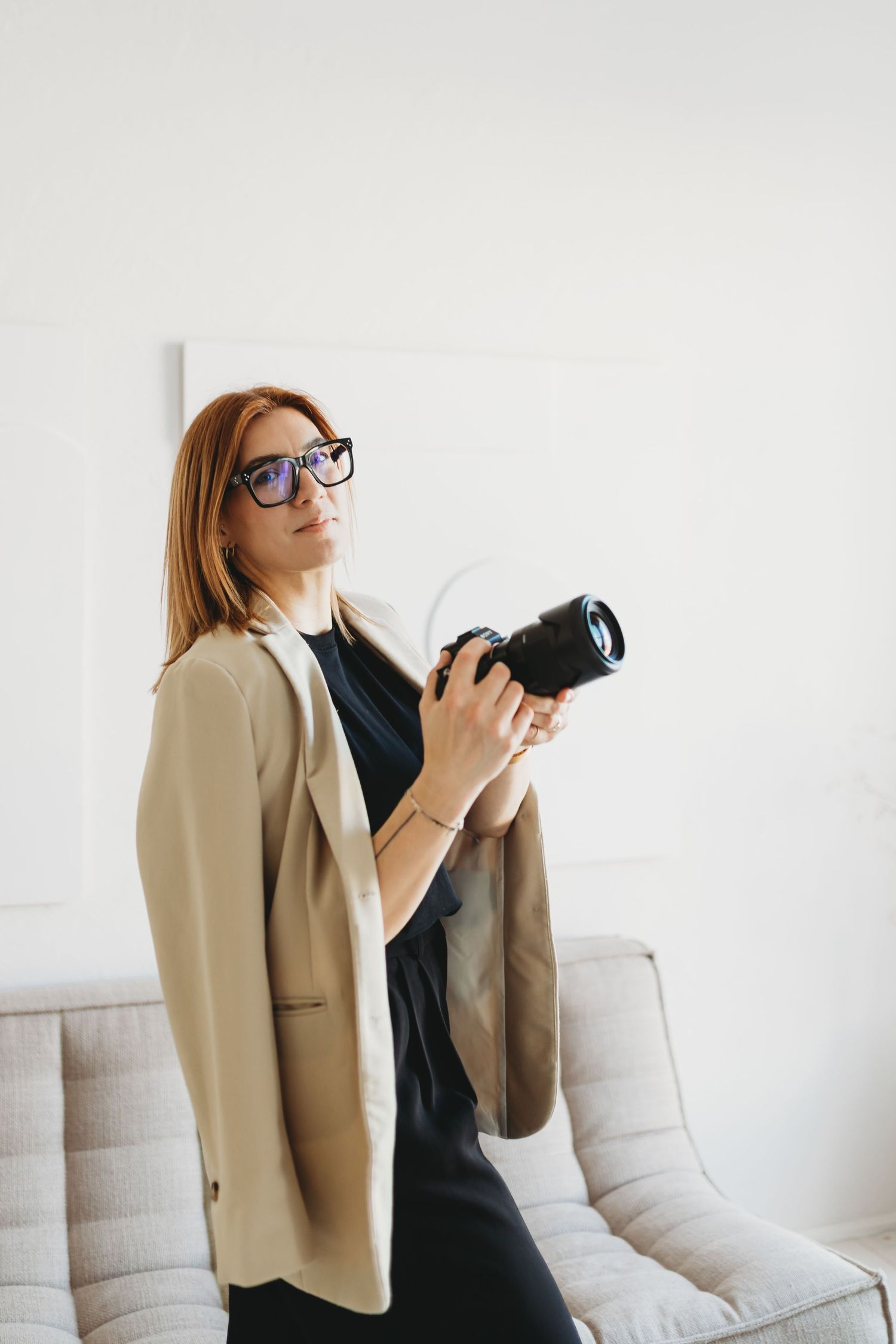 Professional photographer in beige blazer and black dress holding DSLR camera in bright minimal interior setting.