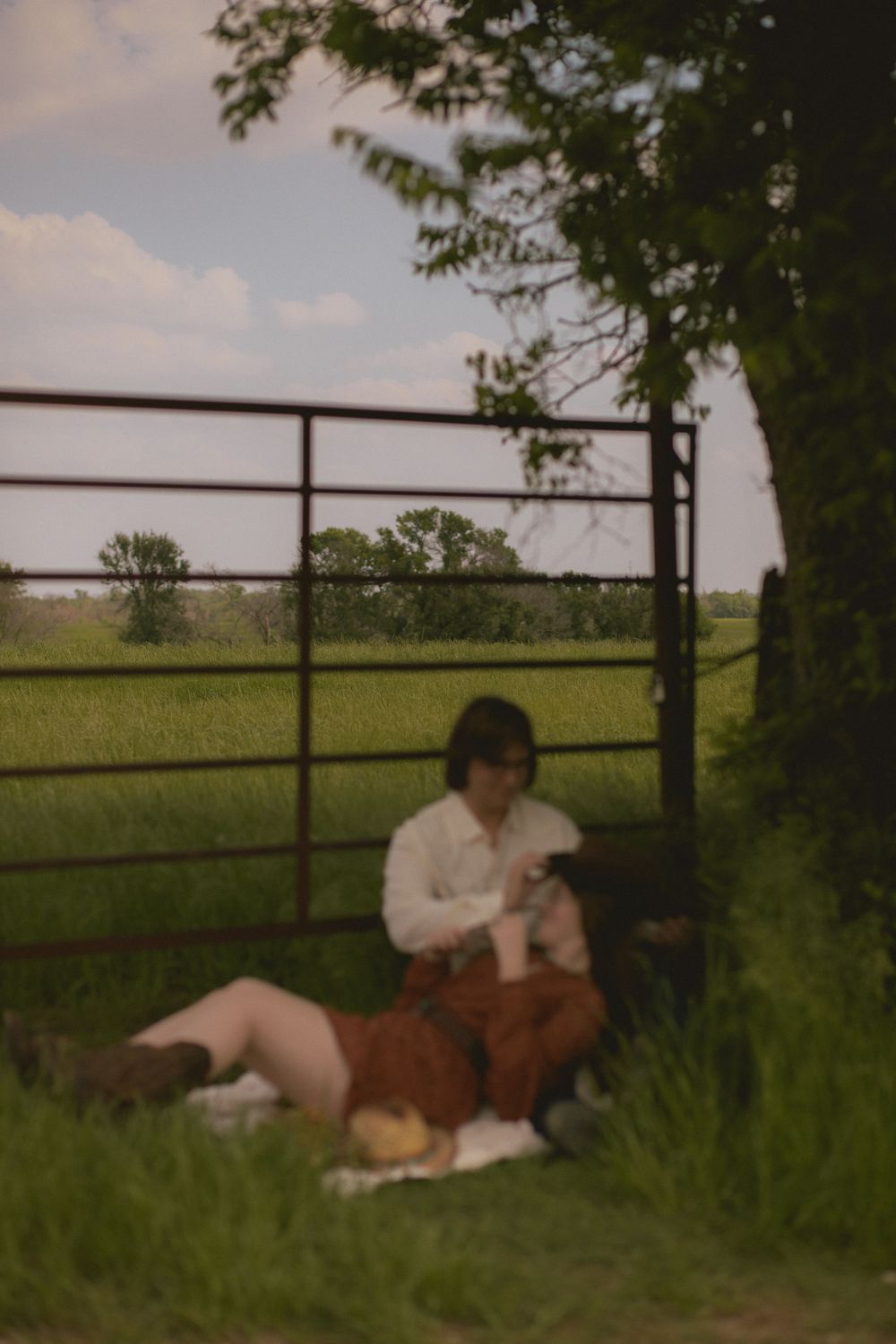 Couple sits together on a blanket by a metal farm gate surrounded by lush green fields.