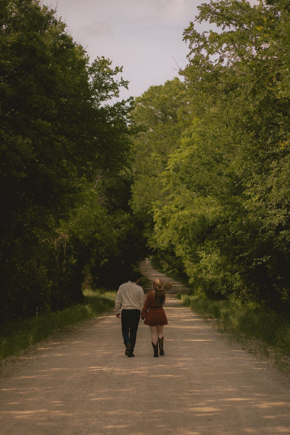 Couple walks hand in hand down a tree-lined dirt path surrounded by lush green foliage.