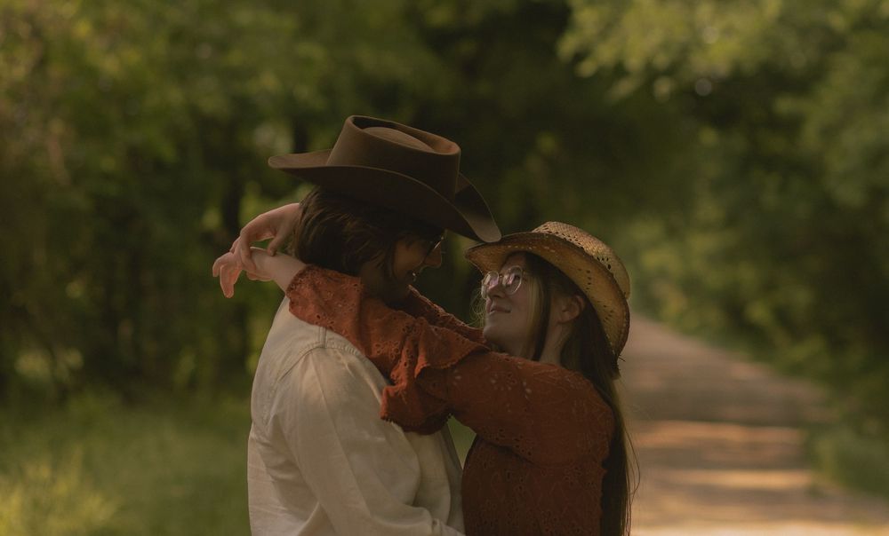 Couple wearing cowboy hats embrace and dance together on a scenic tree-lined country road.