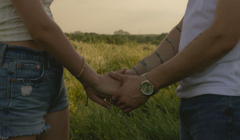 Couple holds hands in a green meadow, man wearing a silver watch, woman in denim shorts.