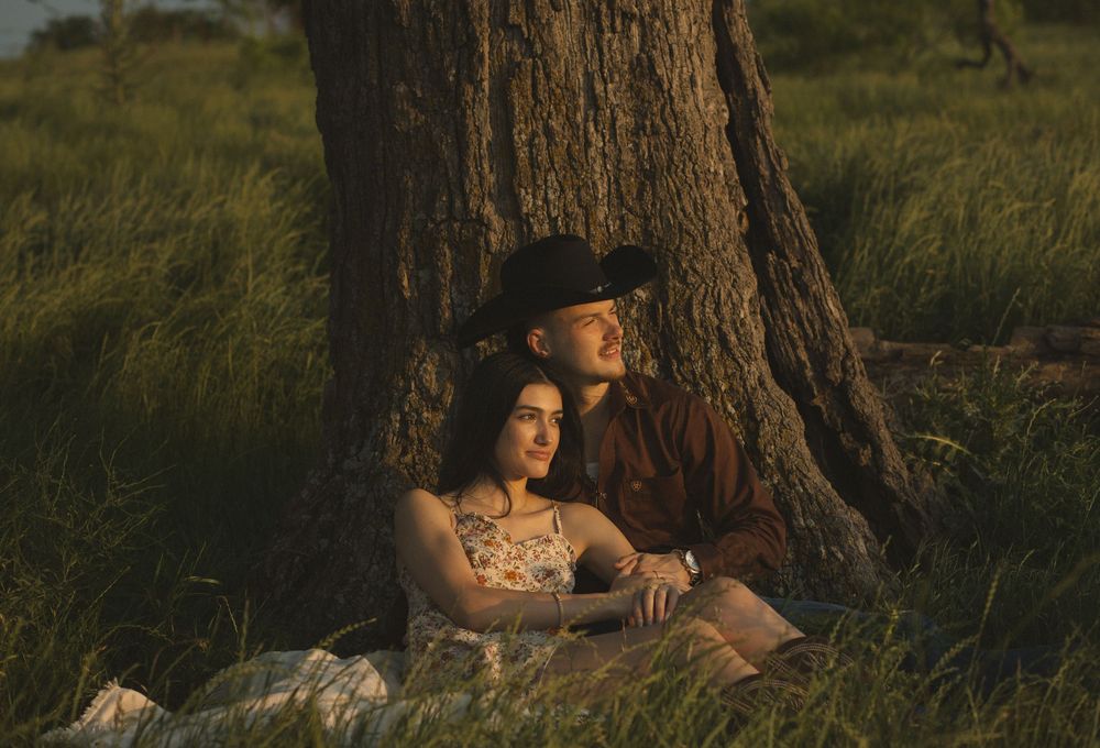 Couple sits leaning against a tree trunk in golden hour light in a lush green field.