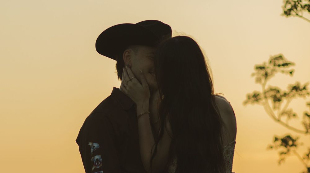 Couple sharing a romantic kiss at golden hour sunset, silhouetted against warm orange sky.