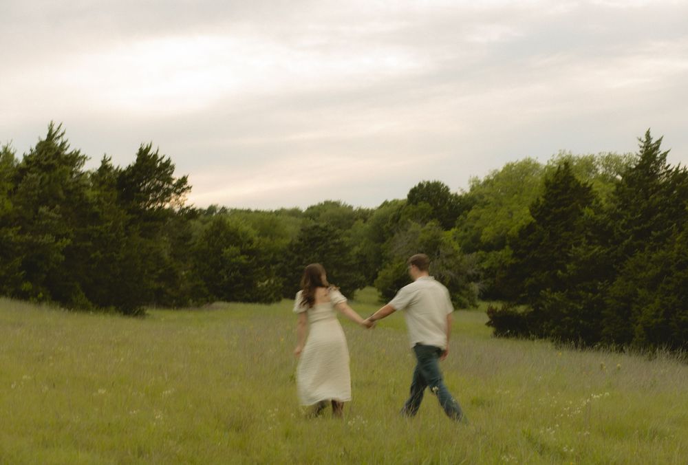 Couple holding hands walking through a lush green meadow at dusk with trees in background.