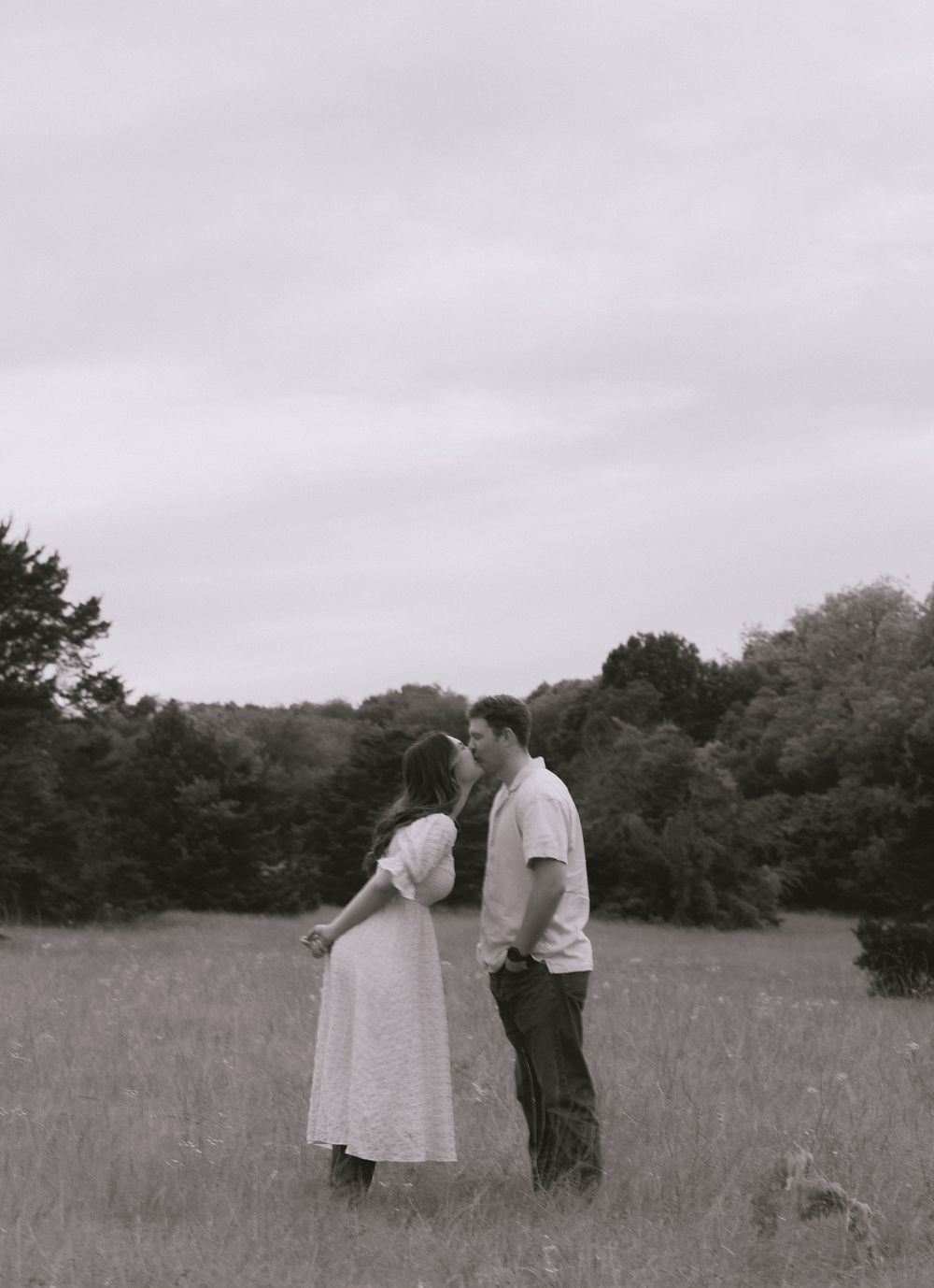 Couple sharing a tender kiss in an open field, captured in black and white with trees behind.