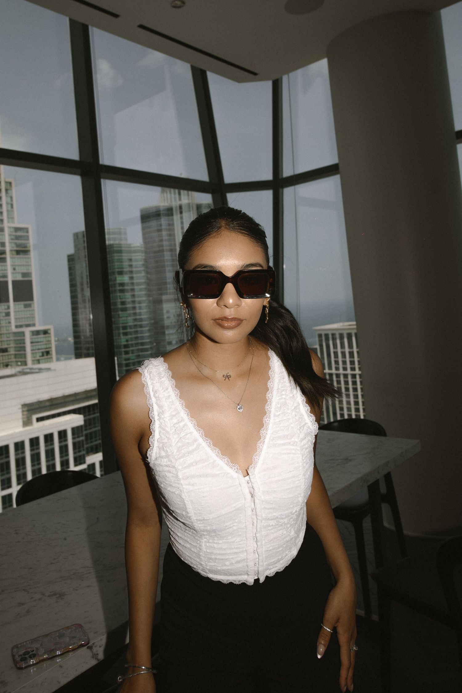 A person in a white sleeveless top poses near large windows in a modern high-rise building interior.