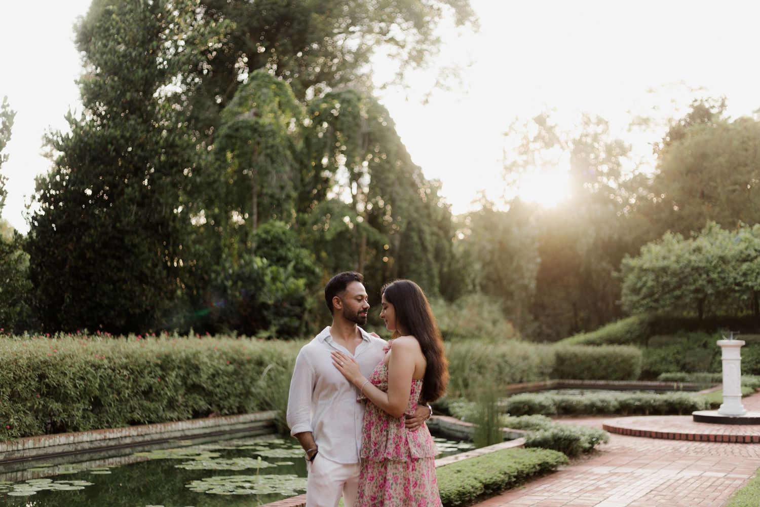 Couple sitting together on grassy hillside during outdoor photoshoot in floral dress and white shirt.