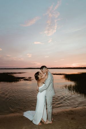 Romantic couple embraces on beach at sunset with water and marshland in background.