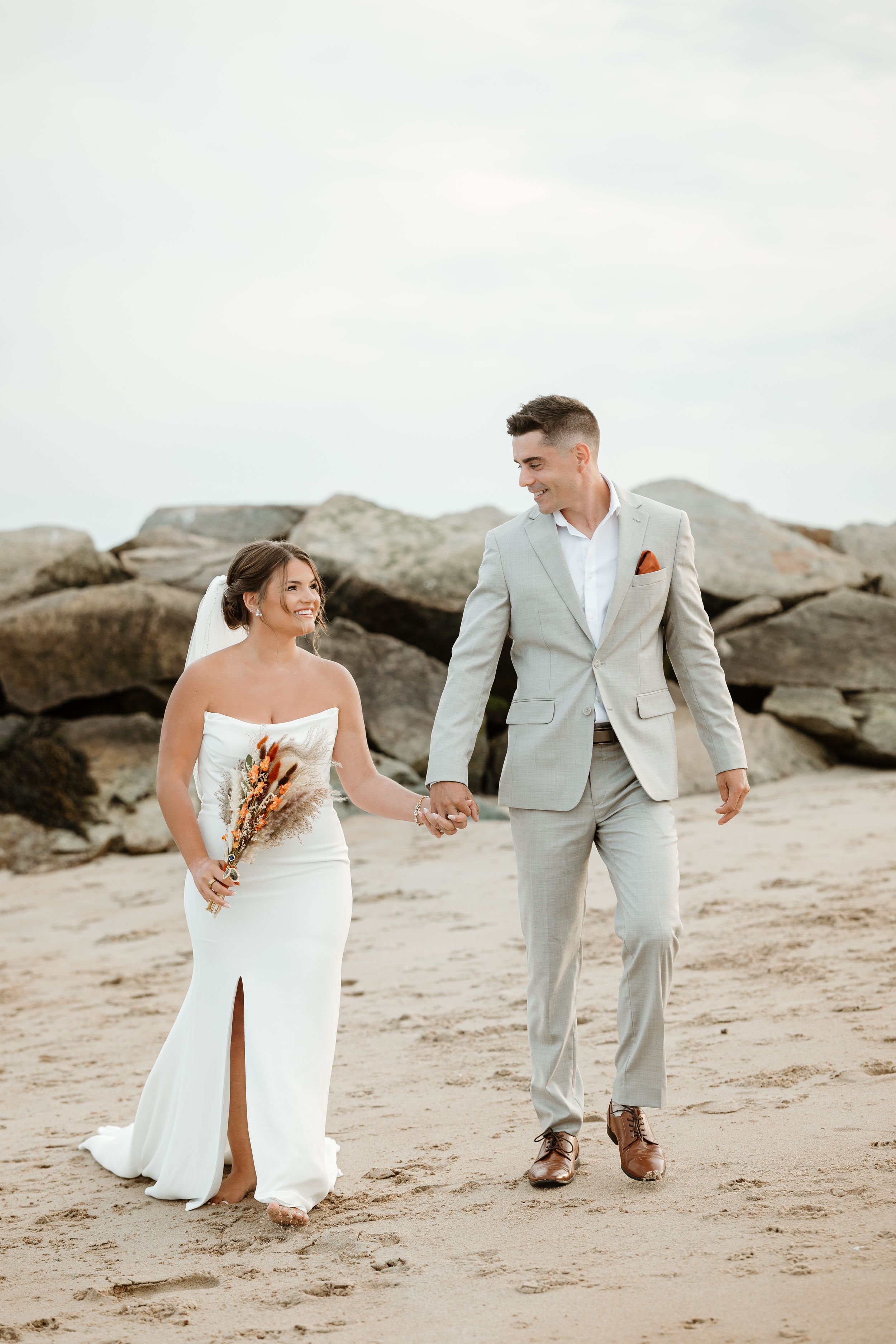 Newlyweds walk hand in hand along rocky beach, bride holding dried flowers.