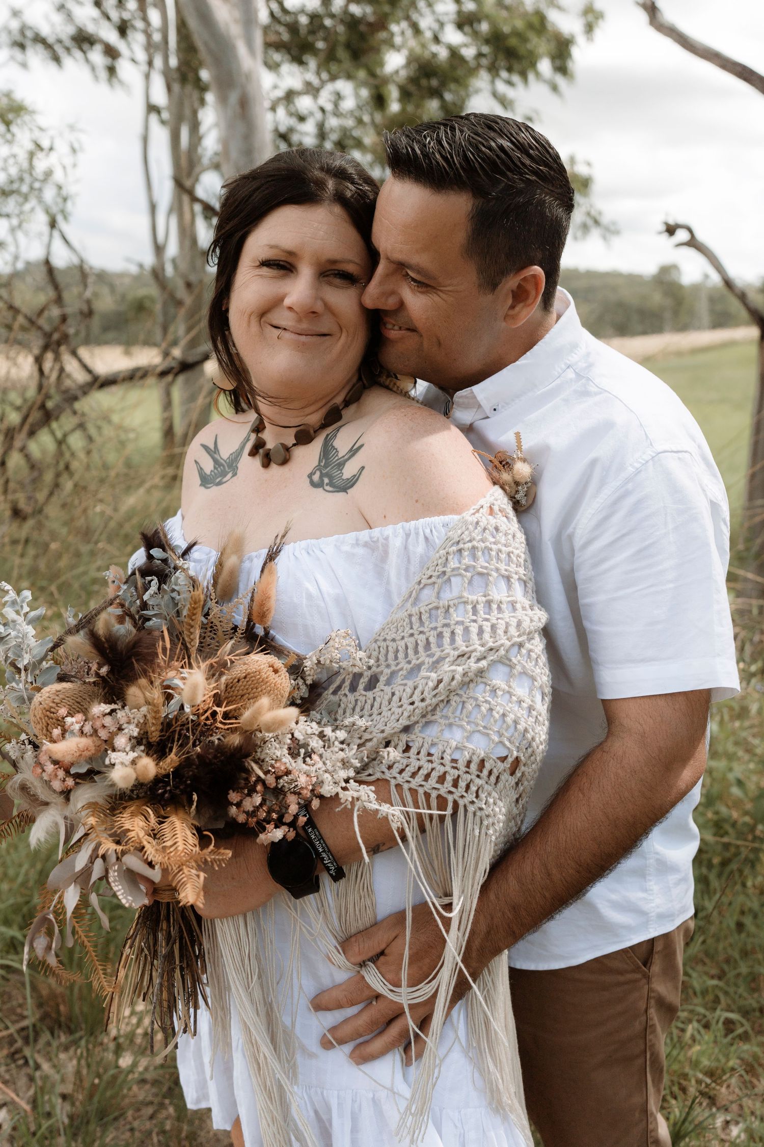 A romantic couple shares intimate moments in a rustic outdoor setting while holding a dried floral bouquet.