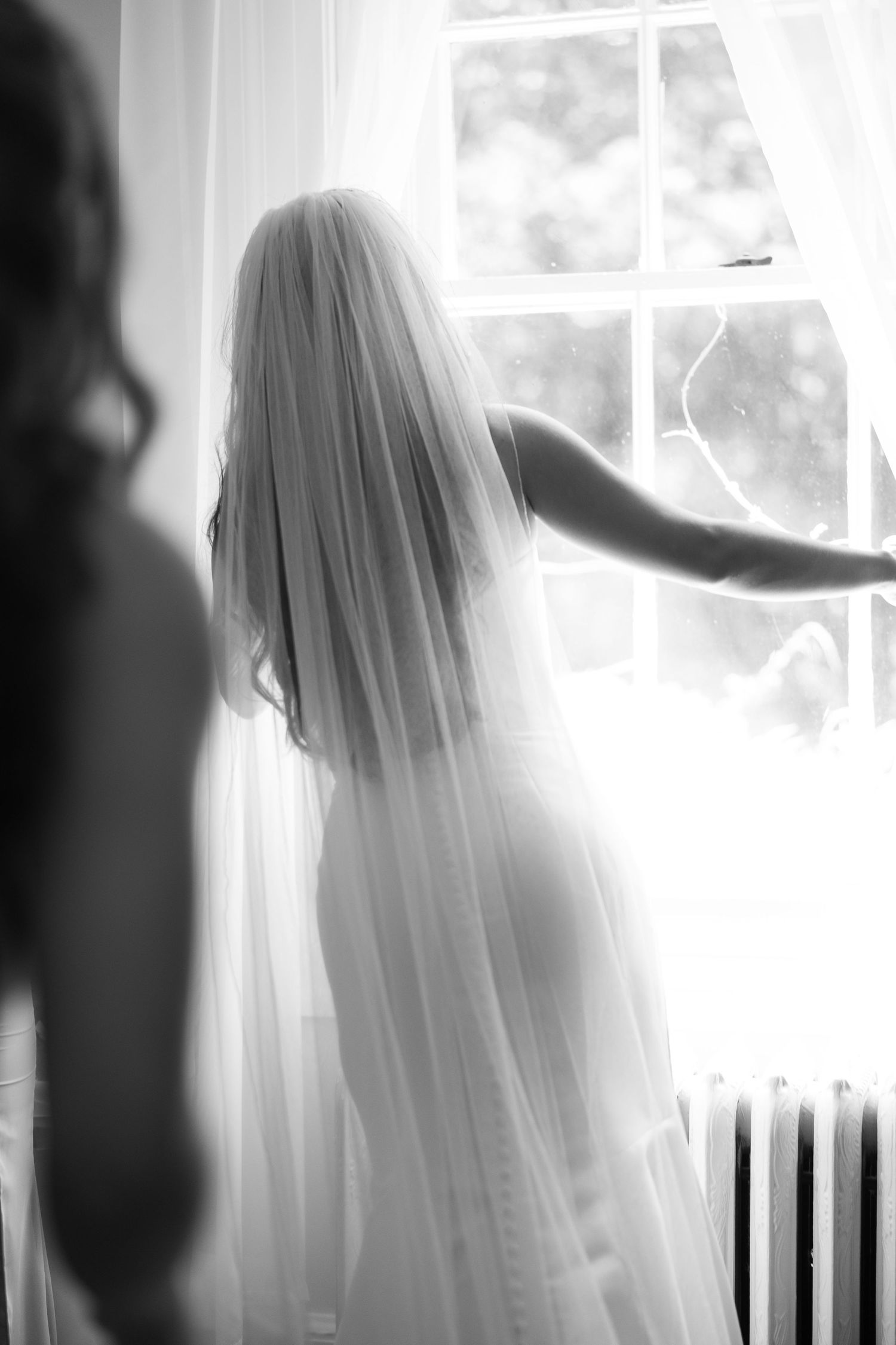 Black and white photo of a bride in a veil standing at a bright window on her wedding day.
