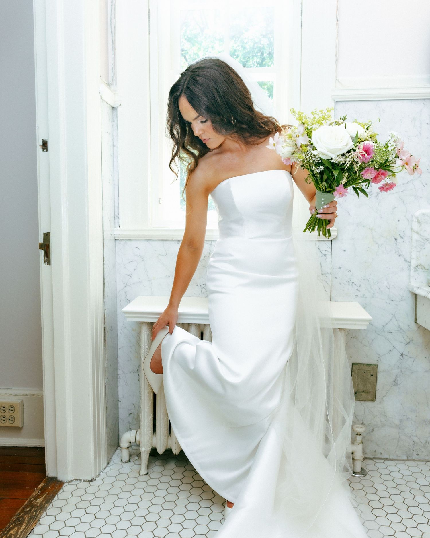 Bride in strapless white satin wedding gown holding pink and white bouquet, standing in elegant marble bathroom.