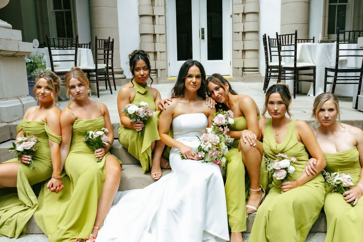 Bride in white gown with bridesmaids in green dresses holding bouquets on stone steps at elegant venue.