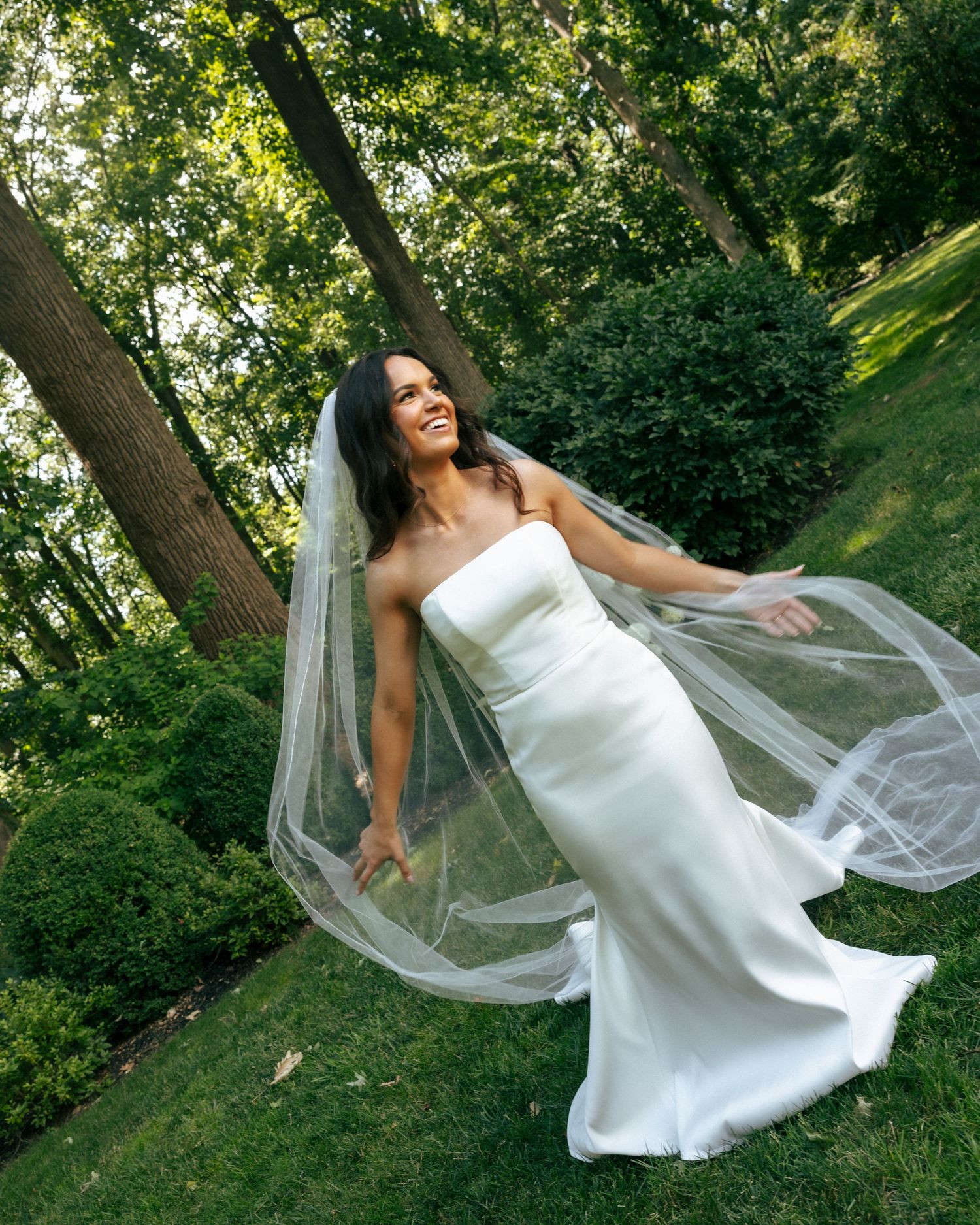 Bride in strapless white gown twirling her flowing veil in a lush green garden setting.