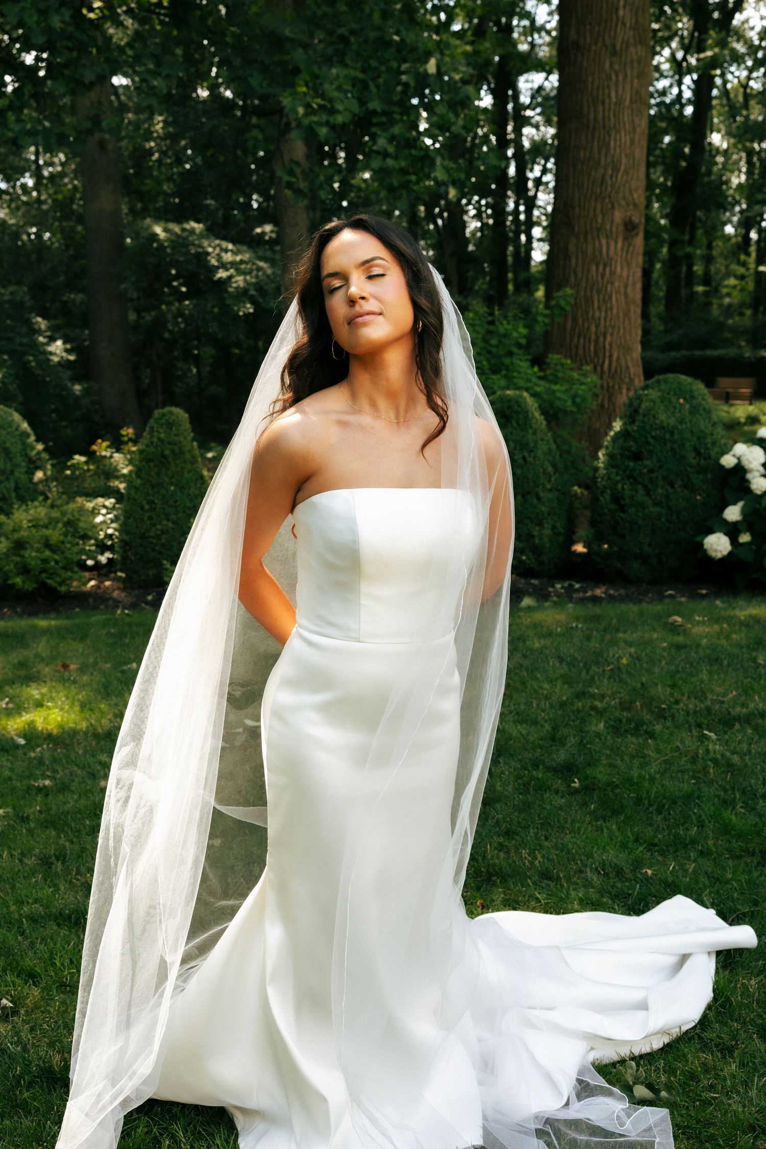 Bride in strapless white gown and flowing veil poses outdoors in lush garden setting.