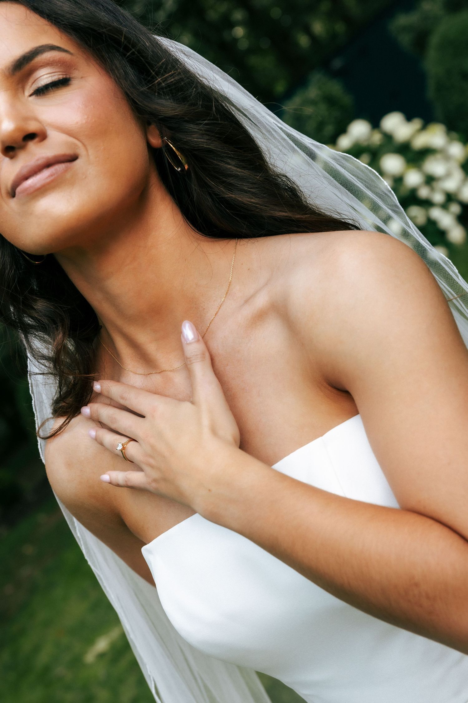 Bride in white strapless gown and veil, eyes closed, hand on chest, wearing gold ring and delicate necklace outdoors.