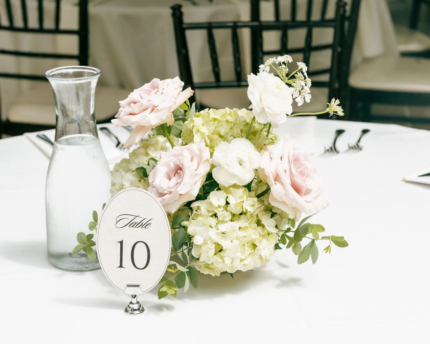 Elegant wedding centerpiece with blush pink roses and white hydrangeas beside a table number 10 sign on a white tablecloth.
