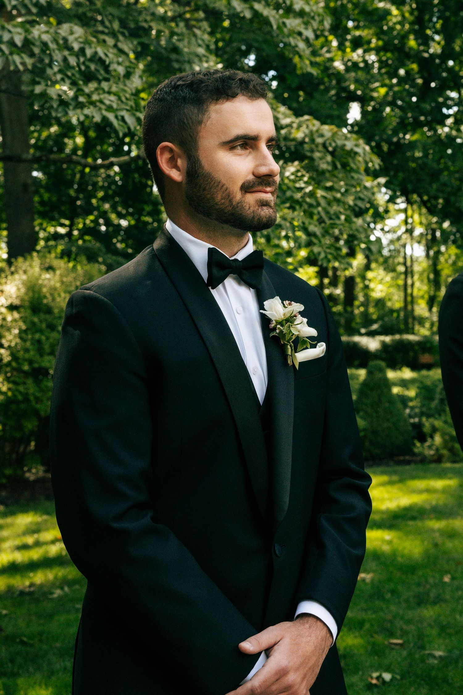 Groom in black tuxedo with white boutonniere standing outdoors in lush green garden.